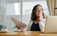 Concentrated online learner in glasses sitting at desk using laptop and reviewing her notes.