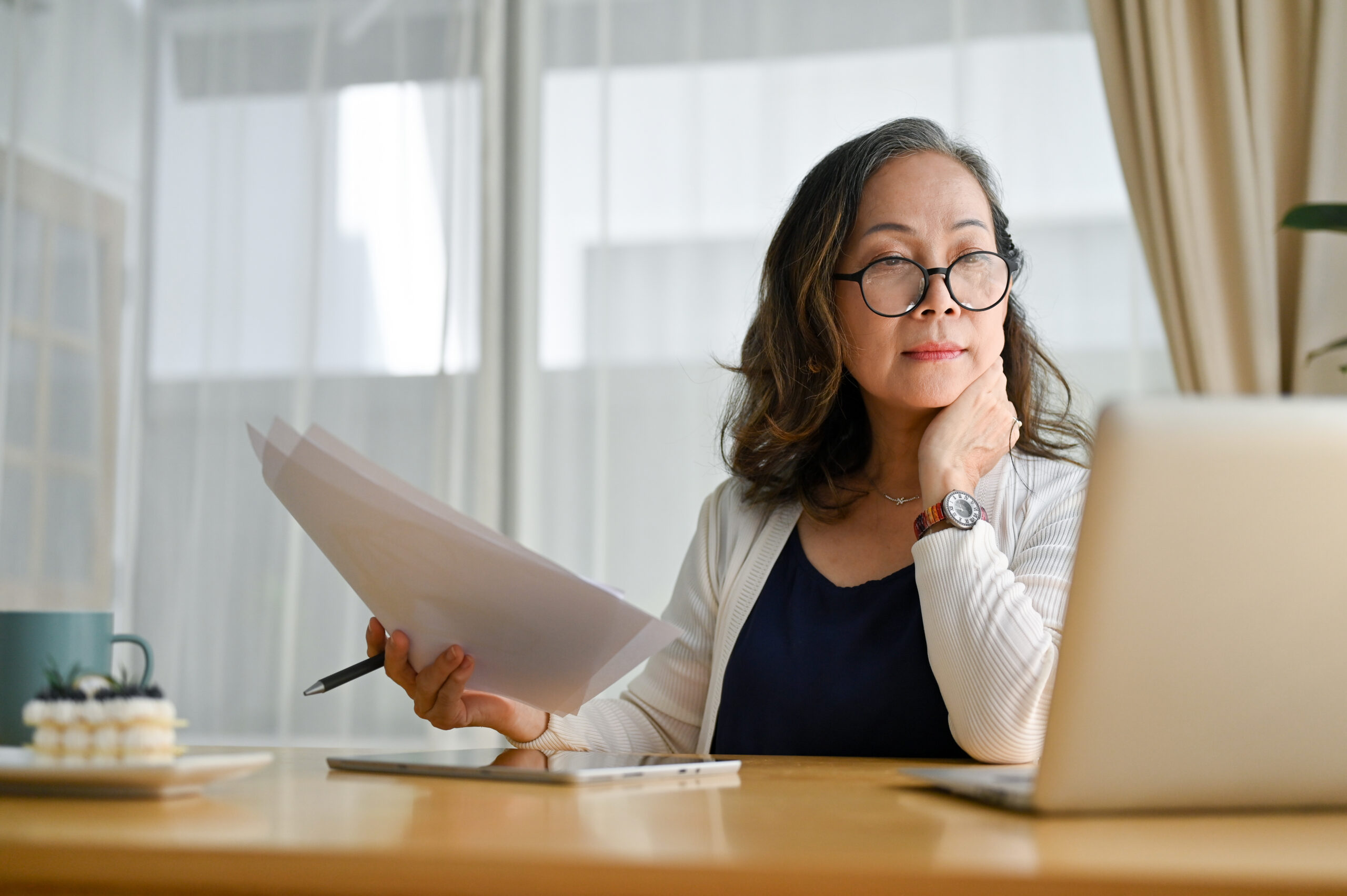 Concentrated online learner in glasses sitting at desk using laptop and reviewing her notes.